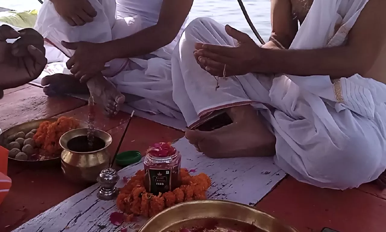 A Hindu religious ceremony (puja) is being performed by men on a boat, with traditional offerings like flowers and sweets placed before them.- Asthi Visarjan in Prayagraj from Singapore